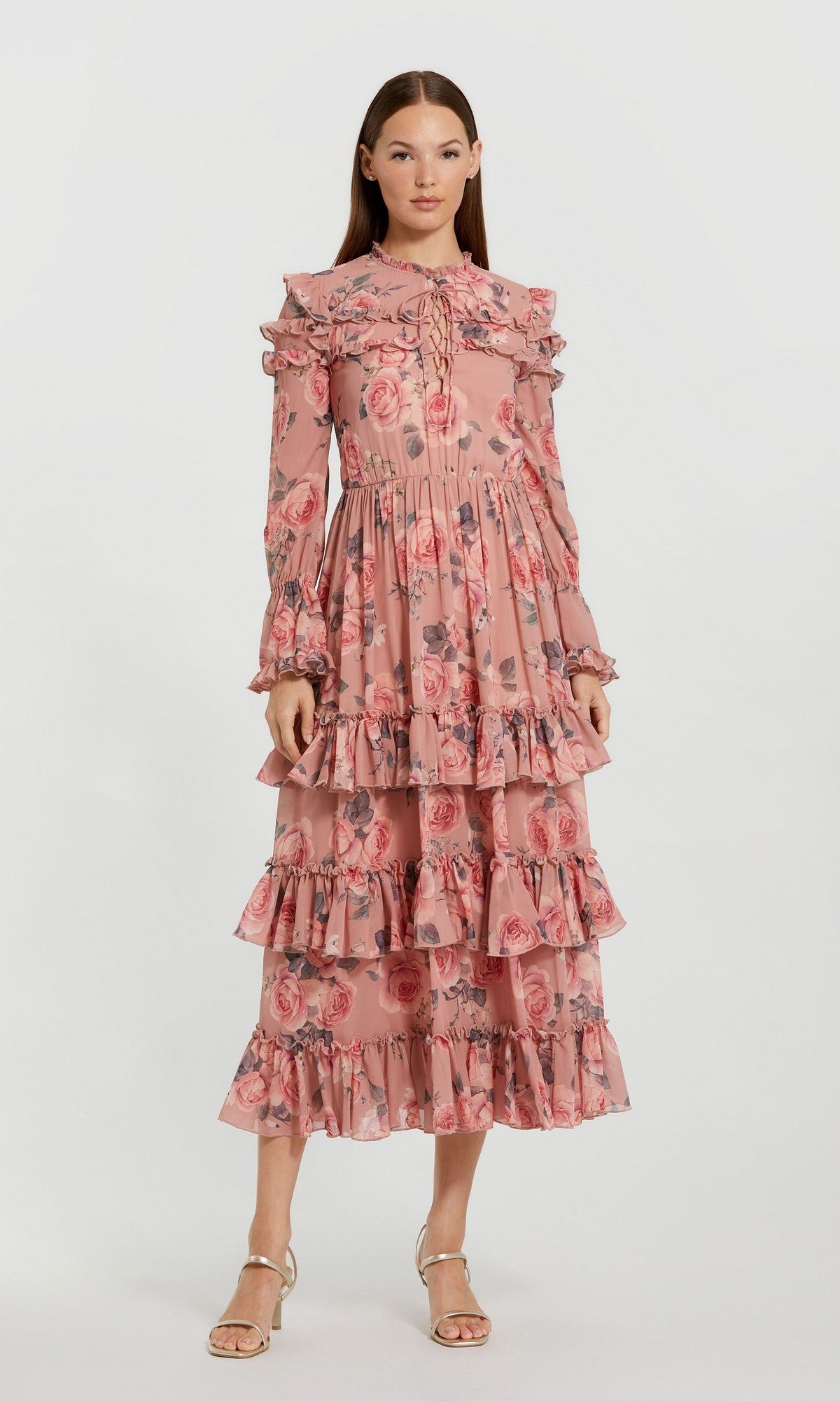 A woman wearing the Midi Wedding Guest Dress: Mac Duggal 9306 in Rose Multi, a long-sleeved chiffon dress featuring pink floral patterns, ruffles, pleats, and a tiered skirt, stands against a plain white background.